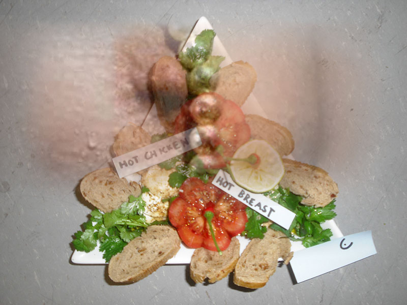 Tomate, Mozzarella, Chili, Brot auf dreieckigem Teller, Text auf Papierstreifen: 'hot chicken, hot breast'; überlagert mit dem Foto einer Brustwarze
